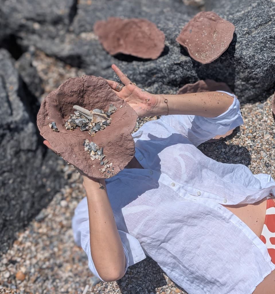Artist on a shell-strewn shore holding a handmade ceramic bowl