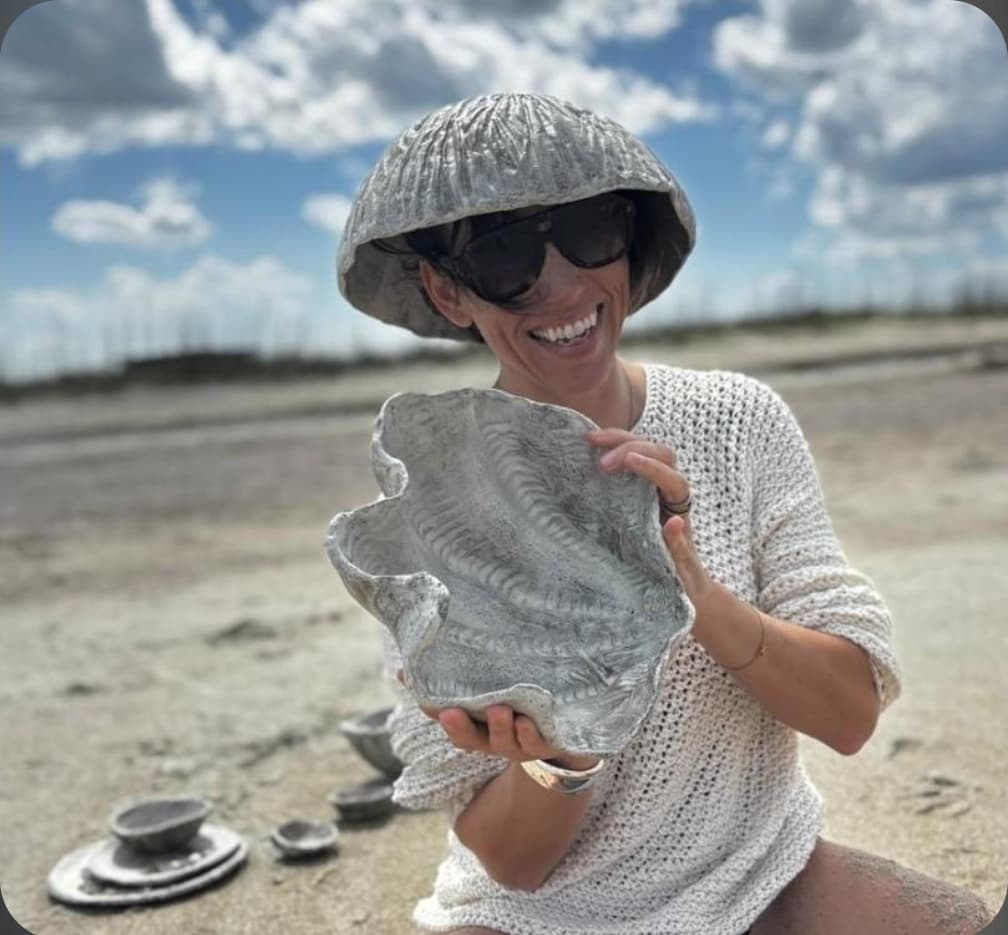 Kimberly on a sandy beach wearing a textured ceramic bowl as a hat and holding a large shell-shaped vessel